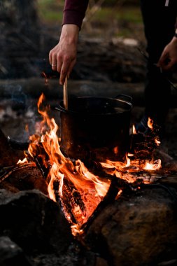 Kaşık tutan ve yanan ateşin üzerinde duran tencerede yemek karıştıran adamlar.