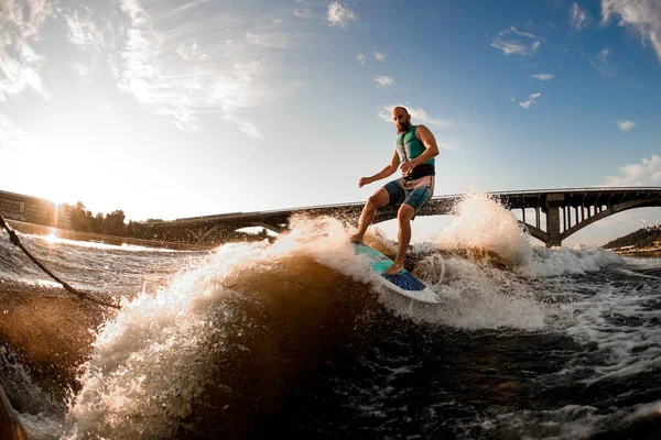 Wakesurfer jadący na wakeboardzie po falach rzecznych przeciwko błękitnemu niebu i mostowi — Zdjęcie stockowe