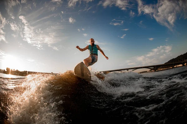 Samiec budzący się na wakeboardzie w dół fal rzecznych przeciwko błękitnemu niebu i mostowi — Zdjęcie stockowe