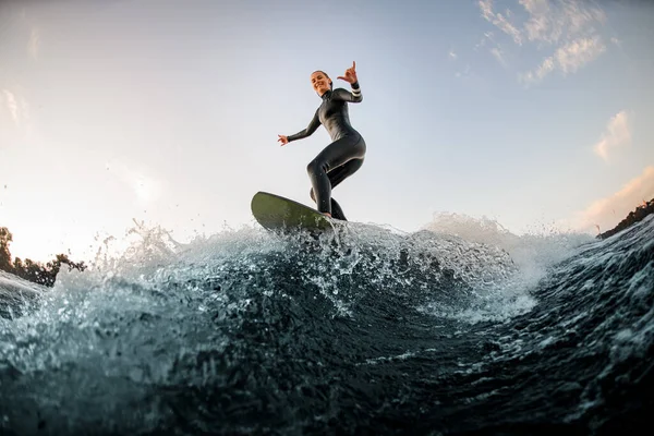 Mulher feliz em wetsuit wakesurfing na placa e andando pela onda do rio e mostrar gesto mão — Fotografia de Stock