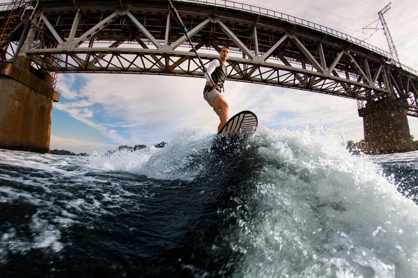 Homme actif tient la corde et monte vague sur planche de wakeboard sur le fond du pont — Photo
