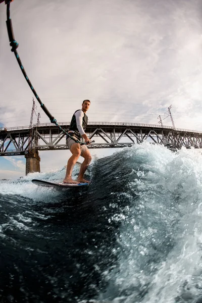 Adulto homem detém corda e cavalga onda no wakeboard bordo contra o fundo da ponte — Fotografia de Stock