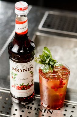 UKRAINE, KYIV - 12 Mart 2021: Monin kızılcık şurubu şişesinin açısı ve bar tezgahında bir bardak ferahlatıcı kokteyl