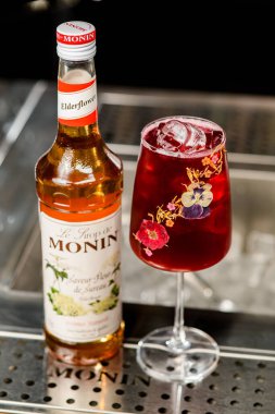 UKRAINE, KYIV - 12 Mart 2021: Saveur fleur de sureau şurubu şişesinin açısı ve bar tezgahında bir bardak soğuk kırmızı kokteyl