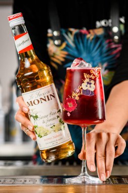 UKRAINE, KYIV - 12 Mart 2021: Barmen el bar tezgahında bir şişe Saveur Fleur de Sureau şurubu ve bir bardak soğuk kırmızı kokteyl tutuyor