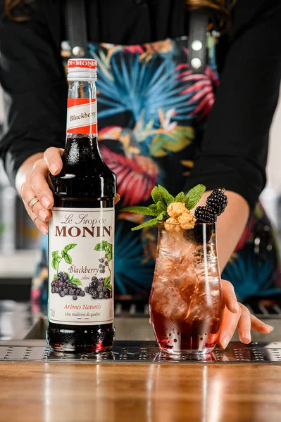 UKRAINE, KYIV - 12 maart 2021: Vooraanzicht van de fles Monin bramen siroop met de hand van de barman erop en glas koude cocktail met bramen — Stockfoto