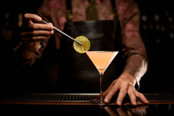 Martini glass with cocktail on bar and hand of bartender decorates the glass with slice of lemon — Stock Photo, Image