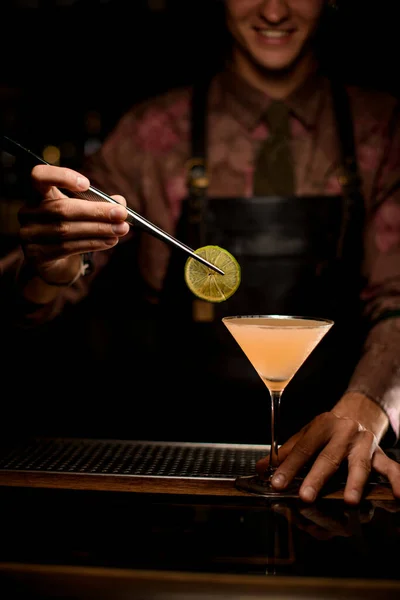 Martini glas met cocktail op de bar en de hand van barman zachtjes versiert het glas met schijfje citroen — Stockfoto