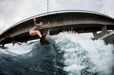 Genç sarışın kadın dalgaların arasında sörf tahtasıyla geziyor. Arkaplanda köprü