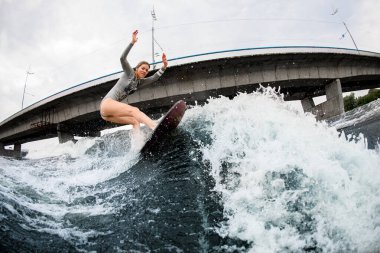 Enerjik bir kadın dalgaların arasında sörf tahtası üzerinde süzülüyor. Arkaplanda köprü
