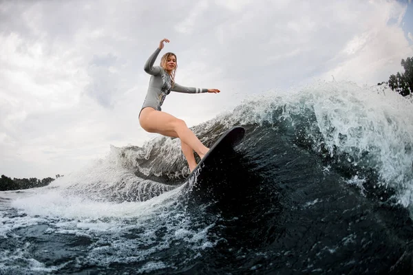 Blonde wet woman in gray wetsuit rides on wakesurf down on huge splashing river wave — Stock Photo, Image