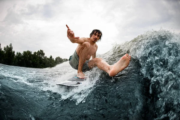 wet man balancing on wake surf board stretching his leg and showing hand gesture