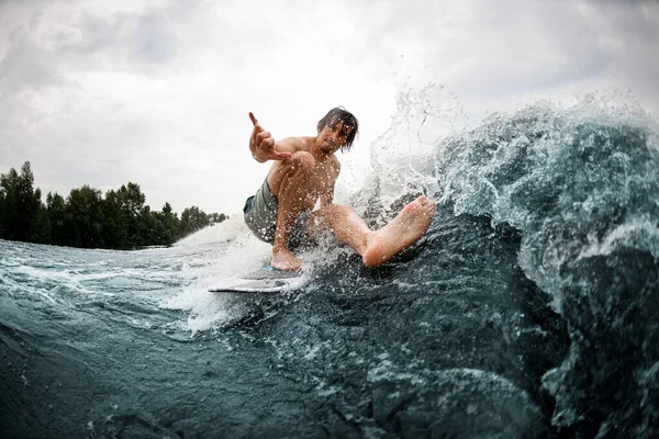 Hombre húmedo activo balanceándose en tabla de surf despertar estirando su pierna y mostrando gesto de mano —  Fotos de Stock
