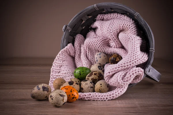 Quail eggs and one orange , green egg  spilling out of basket with pink textile on wooden  background — Stock Photo, Image