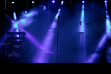 Müzisyenlerin enstrüman ve mikrofonlarının konserden önce dumandan hissedildiği bir konserin ışıkları.