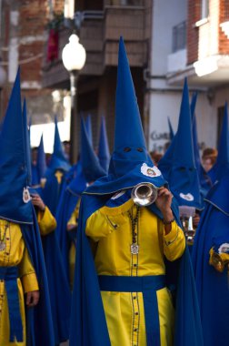 Image of some Nazarenes dressed in robes and hoods with masks participating in a procession playing the music of Holy Week with trumpets and drums