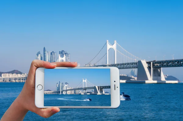 Mano sosteniendo el teléfono inteligente tomar una foto en el puente de Gwangan y Haeundae mar . —  Fotos de Stock