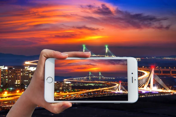 Mano sosteniendo el teléfono inteligente tomar una foto en el puente de Incheon con puesta de sol . —  Fotos de Stock