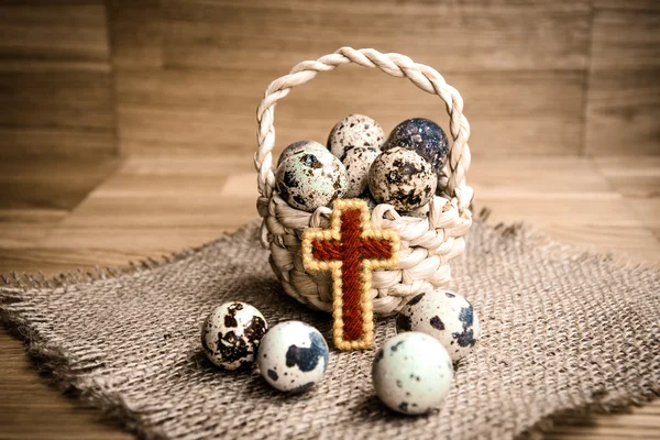 Bird eggs in wicker basket with wooden cross on dark background — Stock Photo, Image