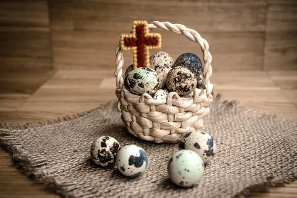 Bird eggs in wicker basket with wooden cross on dark background — Stock Photo, Image