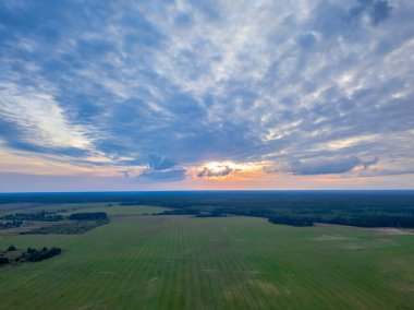 Gün batımında kırsaldaki güzel yeşil bir orman ve tarlanın üzerinde uçmak. Ağaçların ve tarlaların üst manzarası, gün batımında güzel bir gökyüzü. Kuadrokopterin fotoğrafları.