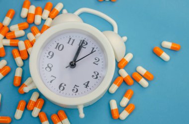 Mavi arka planda beyaz çalar saat ve dağınık hap kapsülleri. Zaman kavramı, sağlık.