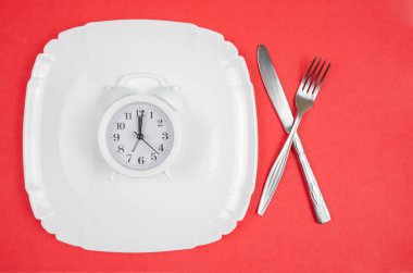 Beyaz alarmlı saat beyaz bir tabakta duruyor. Kırmızı zemin üzerinde çatal ve bıçak var.