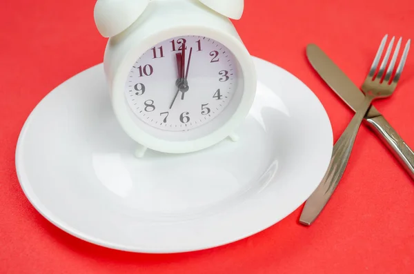 白色闹钟坐在一个白色的盘子上 红色的背景 叉子和刀关闭 晚餐的概念时间 — 图库照片