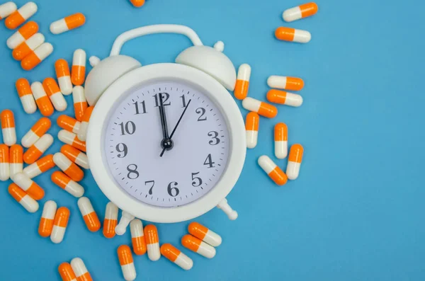 White alarm clock and scattered pill capsules on a blue background close-up. Concept of time, health.