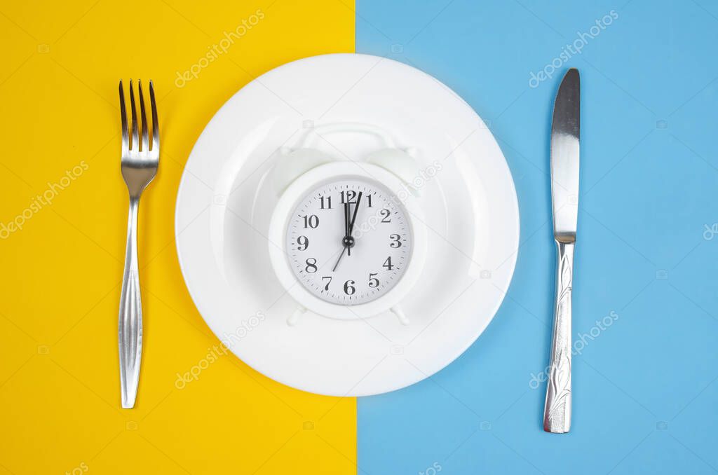white alarm clock lies on a round white plate with a fork and knife on the sides on a blue-yellow background, top view