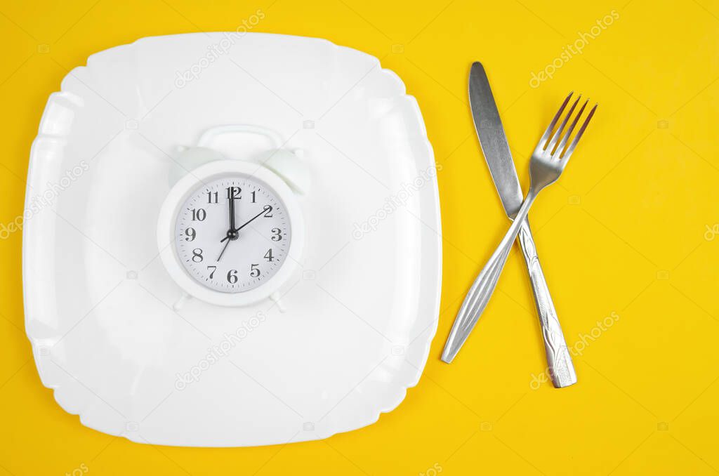 white alarm clock lies on a white plate with a fork and knife on a yellow background, top view