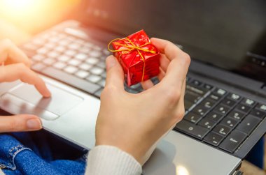 Kadının elinde küçük kırmızı bir hediye kutusu var, yakın plan dizüstü bilgisayarın arka planı, arkadan bir manzara. Aydınlatmalı fotoğraf. Noel kavramı, yeni yıl, tatil