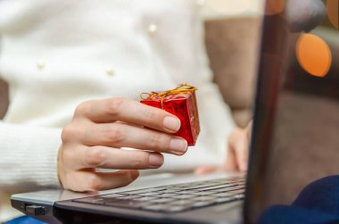 Beyaz kazaklı bir kadın dizüstü bilgisayarını kucağında tutuyor ve elinde kırmızı bir hediye kutusu tutuyor. Noel kavramı, yeni yıl, tatil