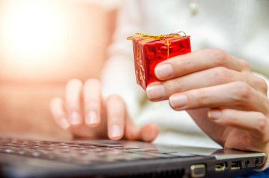 Beyaz kazaklı bir kadın dizüstü bilgisayarını kucağında tutuyor ve elinde kırmızı bir hediye kutusu tutuyor. Aydınlatmalı fotoğraf. Noel kavramı, yeni yıl, tatil