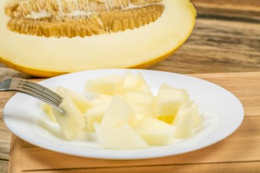 Ahşap bir arka planda beyaz bir tabakta kavun parçaları var, arka planda yarım bir kavun..