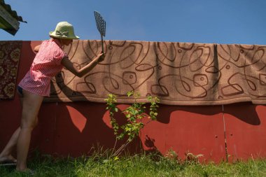 Şapkalı ve gömlekli bir kız güneşli bir günde sokak köşkündeki halıyı temizler, yere serer.