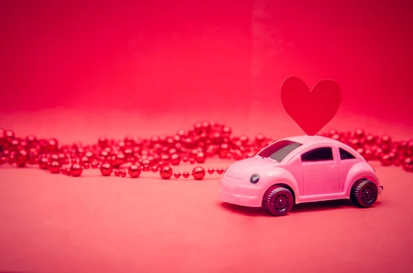 Small pink car with a red heart on the roof of the car on a red background with beads with copy space. Festive bright background