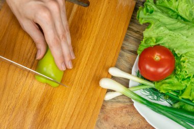 Kadınların elleri, bir tabak sebzenin yanında, tahta bir tahta üzerinde yeşil biber kesiyor. Üst görünüm