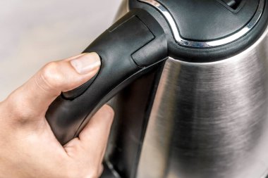 Kadının eli gri bir elektrikli su ısıtıcısının siyah sapını tutuyor ve güç düğmesine yakından basıyor.