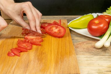 Kadın eli bıçakla kesme tahtasındaki domatesi bir tabak sebzenin yanında kesiyor.