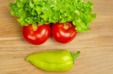 Taze sebzeler, biberler, domatesler, salatalık yaprakları ve su damlaları ahşap bir tahta üzerine güzelce serilmiştir. Sebzeler komik bir yüz şeklinde, üst görünüm.