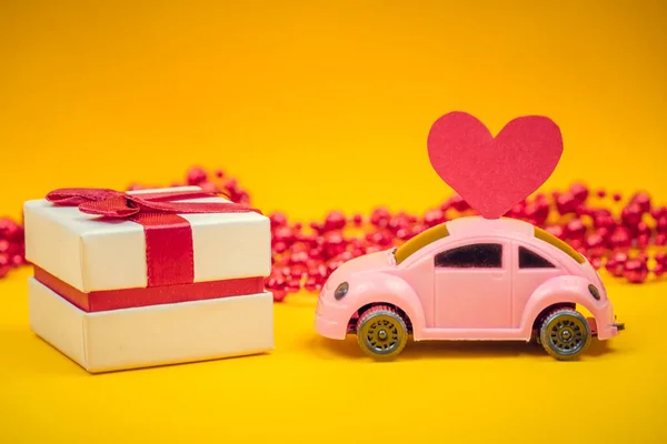 pink toy car with a heart on the roof of the car stands near a gift box and beads on a yellow background with copy space. Holiday background