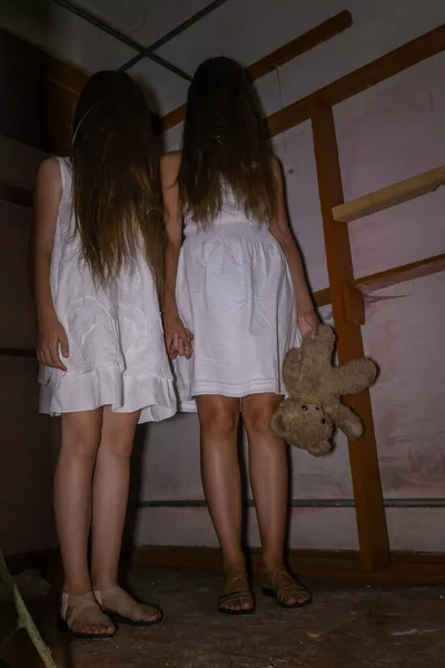 長い髪をした白いドレスを着た2人の女の子テディベアを手に持って暗い放棄された部屋に立って手を握ります 恐怖や神秘主義の概念です 垂直写真 — ストック写真