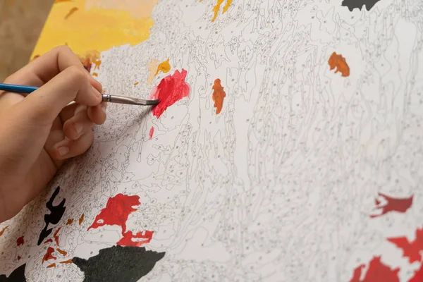 child\'s hand paints a picture by numbers close up with a red paint brush