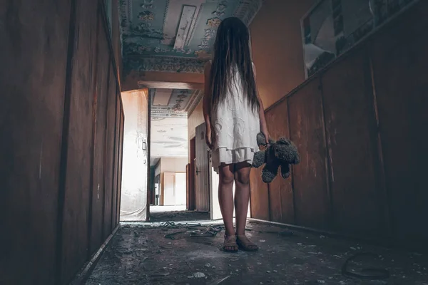 長い髪の白いドレスの少女と手にテディベアが放棄された建物の不気味な廊下の真ん中に立っています 恐怖や神秘主義の概念 — ストック写真