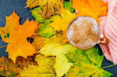 Bir fincan kapuçino kahvesi akçaağaç yapraklarının üzerinde turuncu bir eşarbın yanında duruyor. Sonbahar havası, aile tatili, şükran günü, hafta sonu tatili. Güzel sonbahar arkaplanı