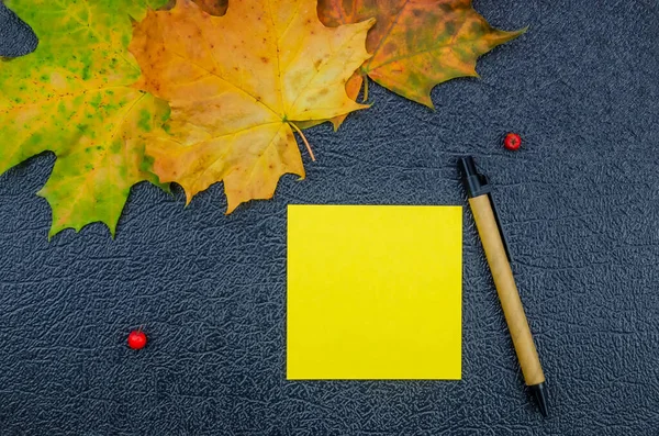 Yellow note sheet, pen surrounded by yellow maple leaves, Rowan berries on a dark background, copy space. Beautiful autumn background. Concept of recording cases and notes.
