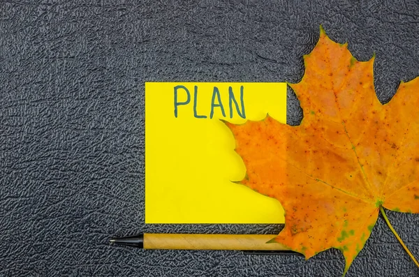 Yellow note sheet labeled Plan with maple leaf and pen on a dark background with space for text, top view. Beautiful background for notes, top view