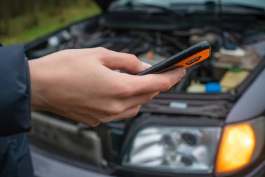 Ceketli bir kadının elinde siyah bir cep telefonu var. Acil bir durakta arabanın kaputuna yakın plan çekilmiş.