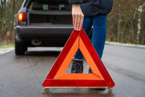 young woman in a winter down jacket in a yellow vest puts an emergency stop sign on the road an orange triangle close up near a car with an open trunk